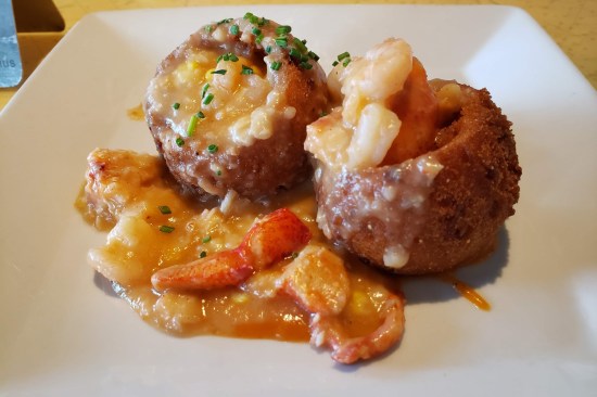 Two large seafood-stuffed hush puppies with buttery lobster broth leaking out the broken tops and onto the plate.