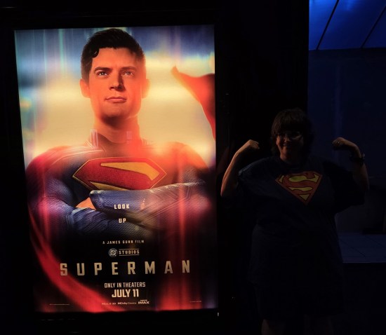 My wife Anne in a blue T-shirt with large Superman S on it, flexing her cute tiny muscles next to a Superman movie poster in a dark theater lobby.