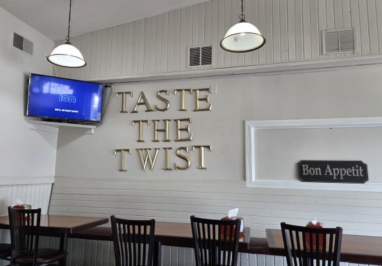 A restaurant dining room with dark wooden chairs, signs saying "Taste the Twist" and "Bon Appetit", and a TV turned to the channel called Ion.