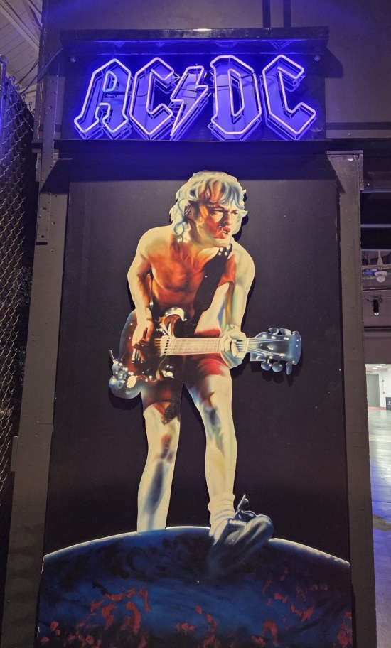 Wall portrait of shirtless young Angus Young playing guitar under a neon AC/DC sign.