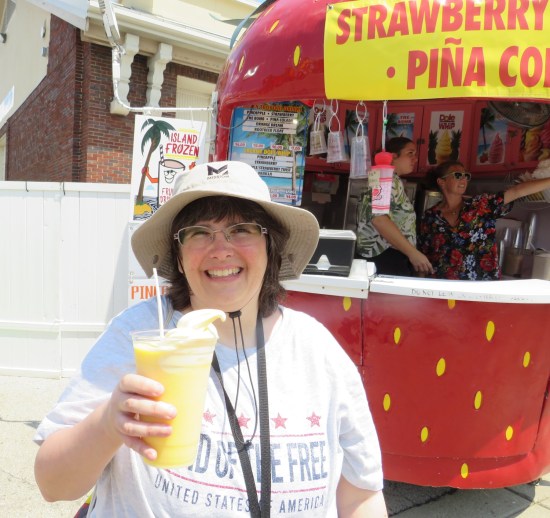 Anne holding a cup with pineapple ice cream in pineapple juice, posing in front of a strawberry-shaped drink stand.