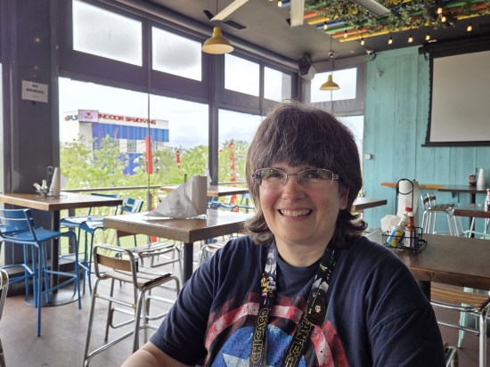 Anne in a restaurant with lots of empty tables and open windows. In the background is a building labeled Indoor Skydiving.