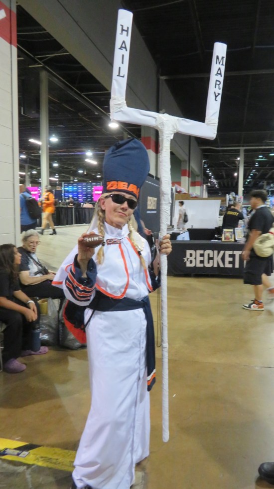 Woman dressed as Catholic clergy with a Chicago Bears mitre, tiny football in one hand, and a white staff shaped like a goalpost, with each prong labeled "HAIL" and "MARY".