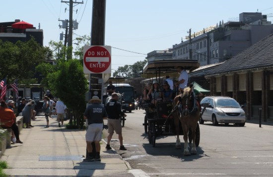 Horse-drawn carriage holding at least a dozen passengers, wheeling down the street near two people in blue polo shirts labeled "Tourism Officer".