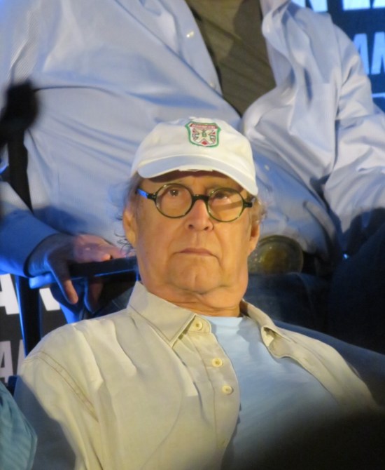 An elderly Chevy Chase with glasses and white cap, glowering directly into the camera.