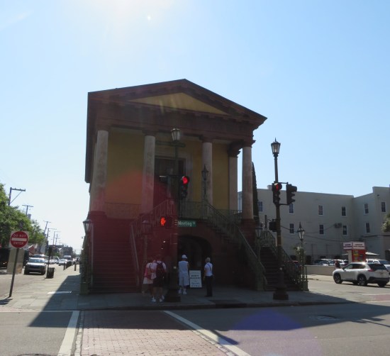 One end of a long, narrow, yellow and brown building in Greek architecture style, with four pillars and a pediment.