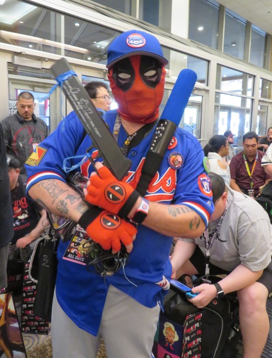 Deadpool cosplayer with Cubs short and hat, wielding a baseball bat and prop gun.