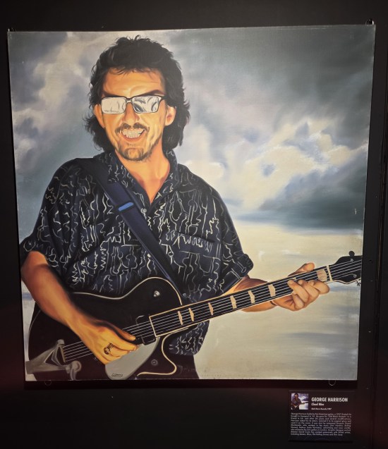 Happy George Harrison with sunglasses and guitar in front of puffy clouds.