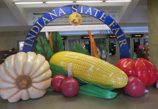 Giant sculptures of corn cob, tomato, carrots, gourd and radishes by an Indiana State Fair arch indoors.