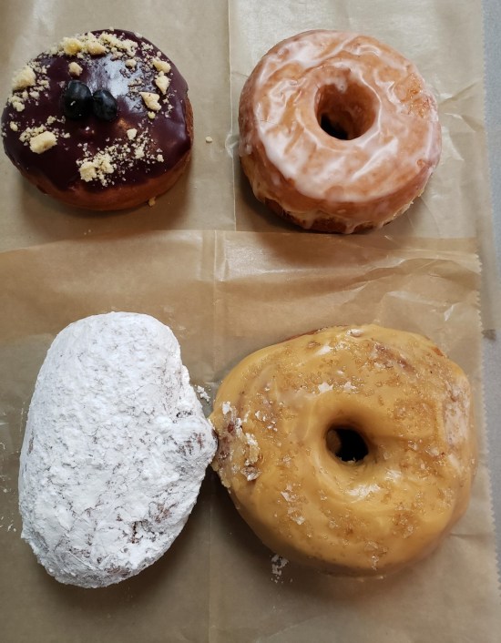 Four donuts on a table. Two are glazed, one is chocolate-covered, one is totally powdered.