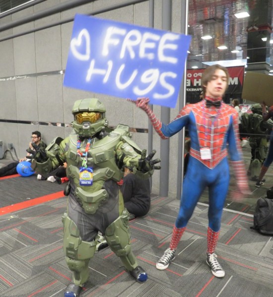 Master Chief cosplayer with arms outstretched, next to a Spidey cosplayer holding a FREE HUGS sign.