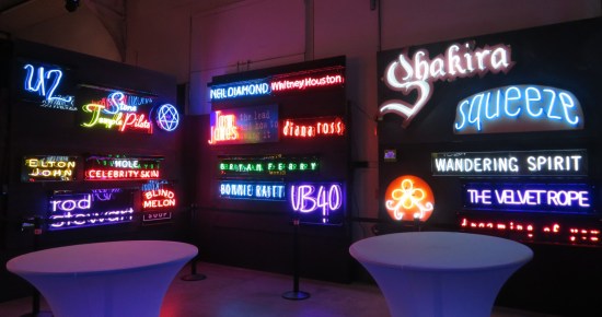 Darkened wall with neon signs of random acts' names including Shakira, U2, Bonnie Raitt, Squeeze, Hole, and more. 