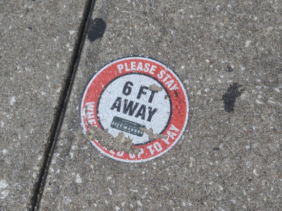 Five-year-old sticker on the street, white circle within a larger red one, reading "Please Stay 6 Feet Away". Another message is eroded and illegible.