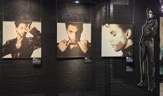 Three different sepia Prince photos hanging on a black-curtained wall next to a Prince standee.