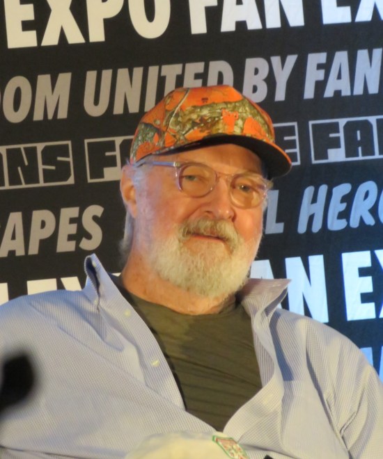 Randy Quaid with glasses, white beard and orange hat.