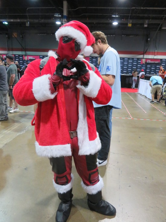 Deadpool cosplayer with Santa jacket and cap, making a heart shape with his hands.