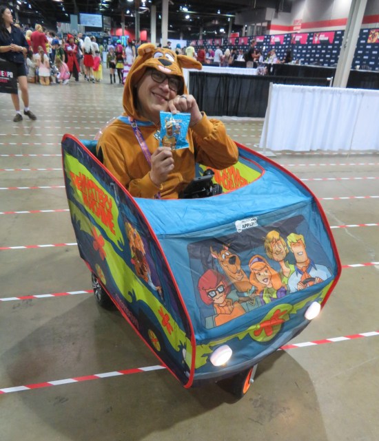 Guy in Scooby-Doo furrry suit, riding in a self-driven Mystery Machine tent-like mini-vehicle and holding up a pack of cinnamon graham Scooby Snacks.
