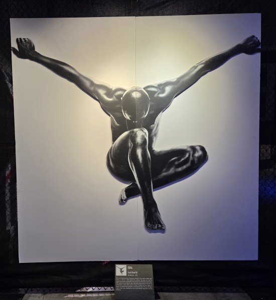 Black-and-white photo of giant black man jumping from somewhere way up high, falling with arms wide and legs crouched for impact.