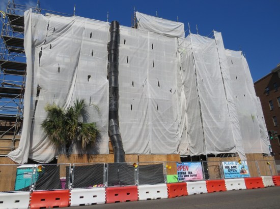 Four-story building or maybe five, covered all in plastic for construction. Businesses on ground floor have signs insisting they're still open.
