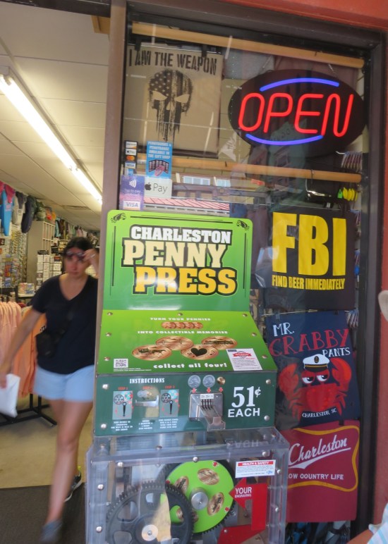 A green smashed penny machine called Charleston Penny Press with price tag of 51 cents. Cheesy souvenir t-shirts hang in the window behind it.