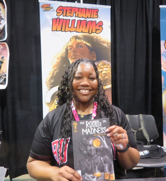 Stephanie Williams at her table holding up a copy of her new project "Roots of Madness".
