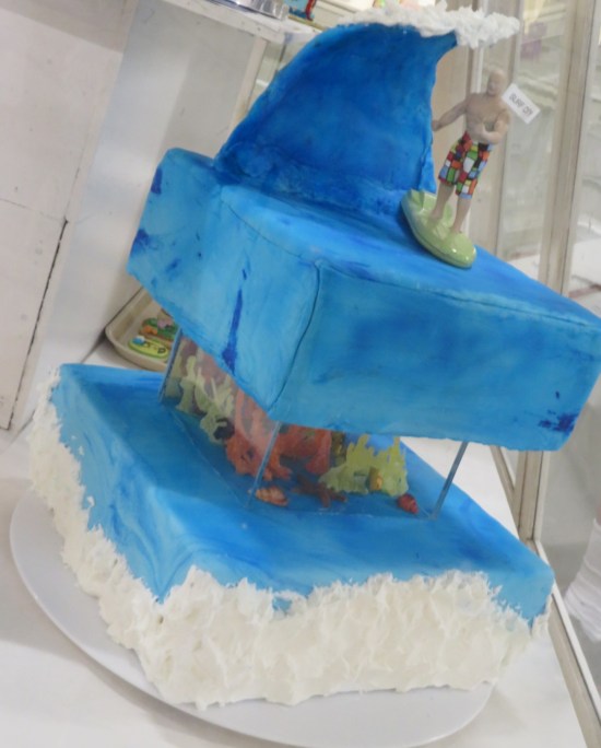 Blue two-tier cake. On top is a big blue icing wave and a surfer figure.