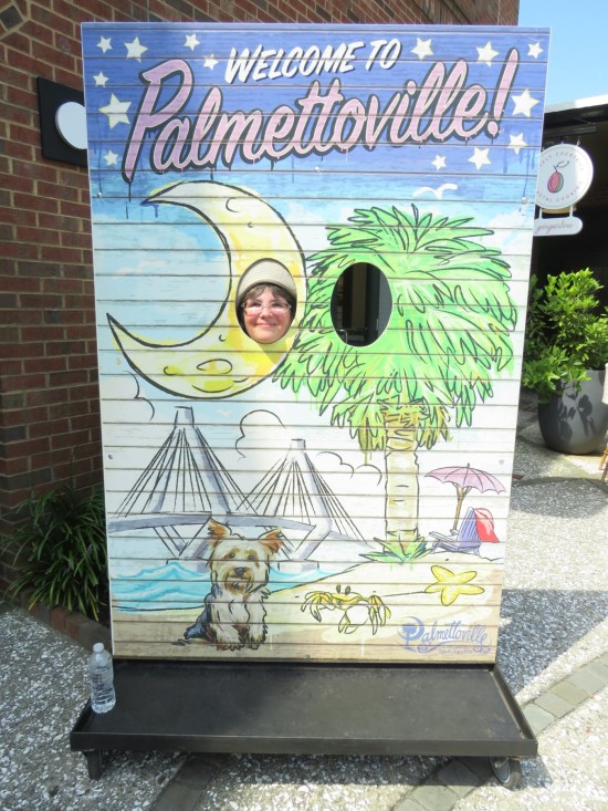 Anne sticking her head through the head-sized hole of a cardboard sign reading WELCOME TO PALMETTOVILLE. The hole is cut so she's sticking her head through a drawing of a crescent moon next to a colossal palmetto and above the Ravenel Bridge and a beach with a doggie.
