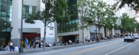 Long line of folks on the sidewalk outside a large building.
