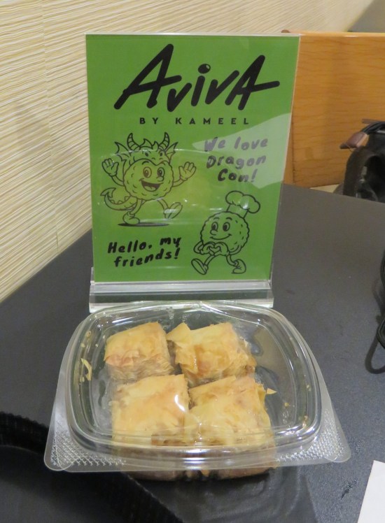 Package of baklava next to an Aviva sign with cartoon falafel balls announcing, "HELLO, MY FRIENDS! WE LOVE DRAGON CON!"