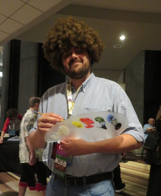 Bob Ross cosplayer with large afro wig, paintbrush and palette.