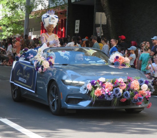 Fancy convertible with flowers in the grill, a Queen Charlotte cosplayer riding on the retracted top, and a Queen Charlotte banner on the side.