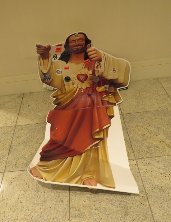Cardboard standee of Buddy Christ from Kevin Smith's Dogma, fallen in a hotel hallway and with not much junk thrown on it.