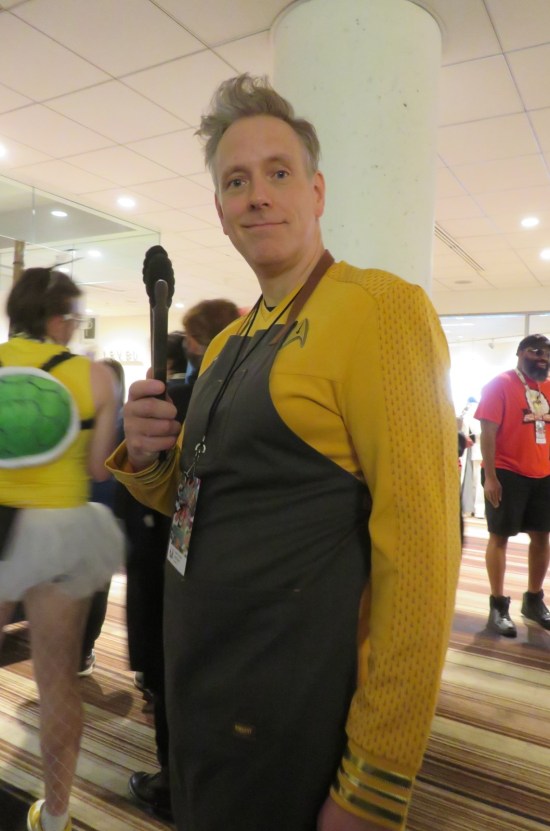 Captain Pike cosplayer wearing an apron and carrying kitchen tongs.