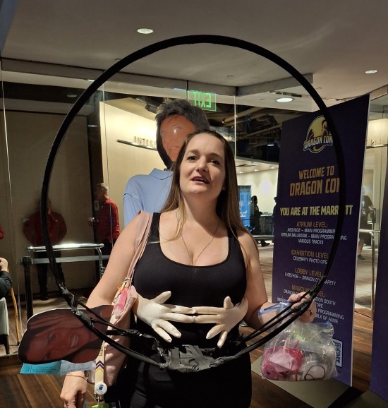 Single cosplayer dressed as Kristin Cabot, holding a hula hoop like a camera frame and with a lo-fi Andy Byron puppet on her back with its arms around her waist.