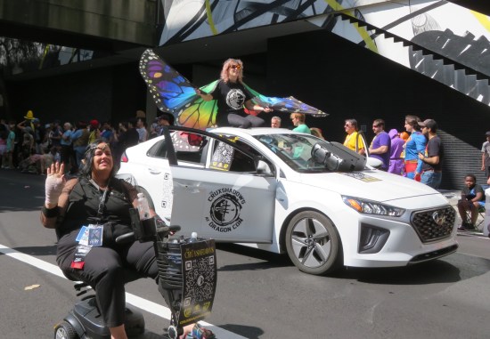 White Hyundai sedan with sunroof Cruxshadows logo on door. Woman with butterfly wings stands inside sunroof; riding alongside is black-clad elf on seated scooter.