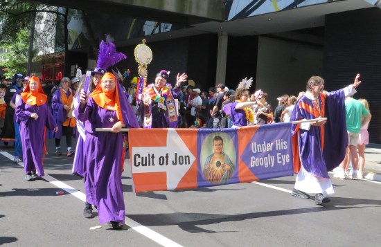 Cosplayers in cult robes colored purple and orange like the Federal Express logo. Their banner has googly eyes on it.