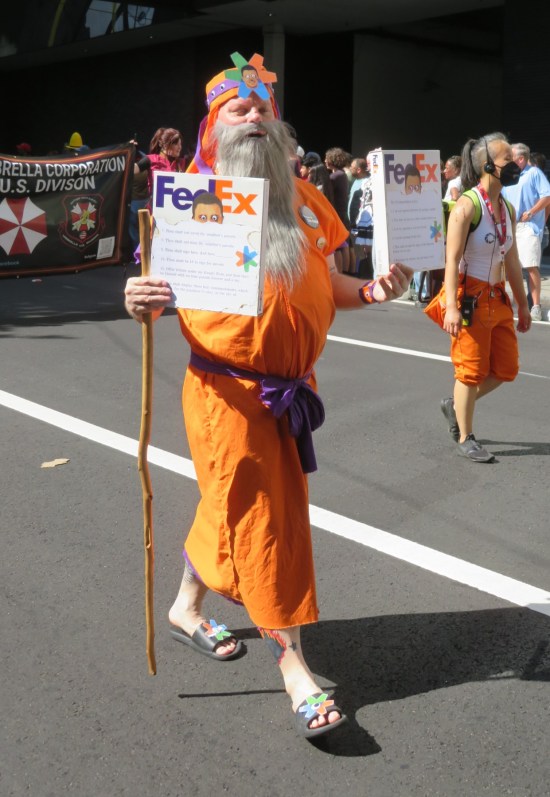 man in orange robe and big fake white beard, carrying a walking stick and two thin Fed Ex boxes.