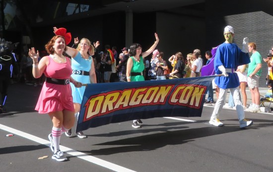 Cosplayers of Mojo Jojo, Blossom, Bubbles and Buttercup walking with a Dragon Con banner.