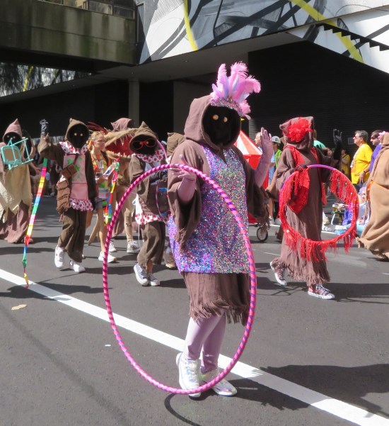 Jawa costume with purple feather headdress, glittery acrobat dress, and carrying a glittery hula hoop. More Jawa are in the next row.