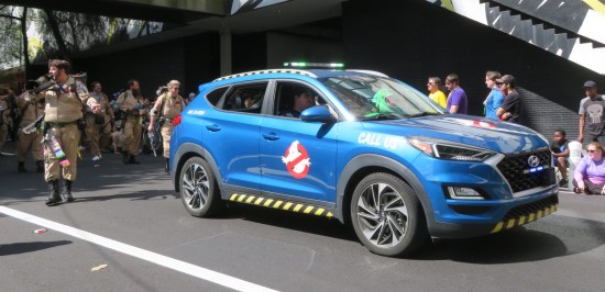 Blue SUV with Ghostbusters logo on door, followed by dozens of walking Ghostbusters.