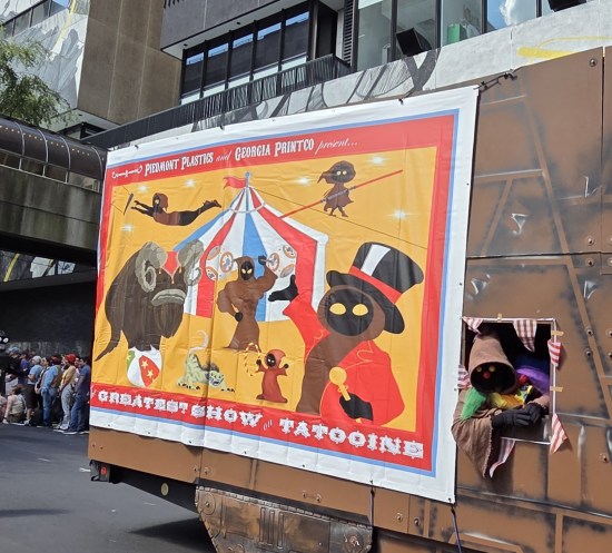 Banner on side of real-life Jawa Sandcrawler, depicting cartoon Jawa holding their own circus. Titled The Greatest Show on Tatooine. A Jawa with clown nose and eyes leans out a window.