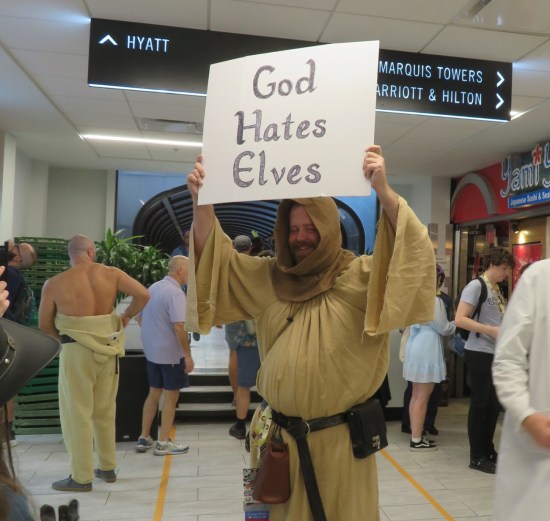 guy in hooded robe holding sign saying "GOD HATES ELVES" standing outside Dairy Queen.