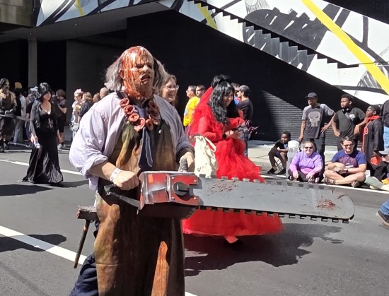 Leatherface cosplayer with huge fake chainsaw, next to Lydia from "Beetlejuice" in her red wedding dress.