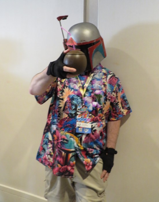 Guy with Boba Fett helmet, gloves and tropical shirt, enjoying a fruity drink served in a thermal detonator.
