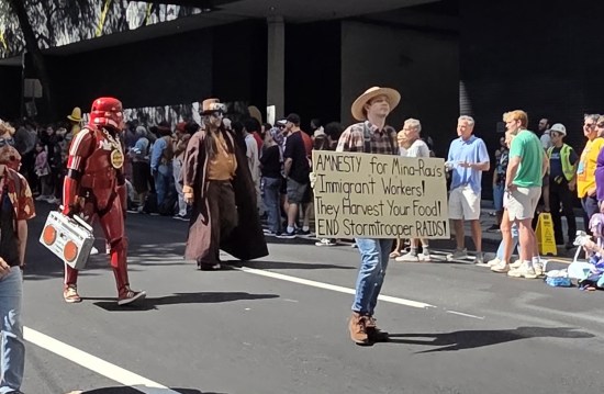 Mina-Rau farmer costume with picket sign: "Amnesty for Mina-Rau's immigrant workers! They harvest your foods! End Stormtrooper raids!"