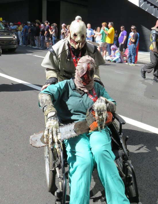 One Netherworld monster cosplayer pushing a Netehrworld serial killer in a wheelchair, who has a chainsaw on their lap.
