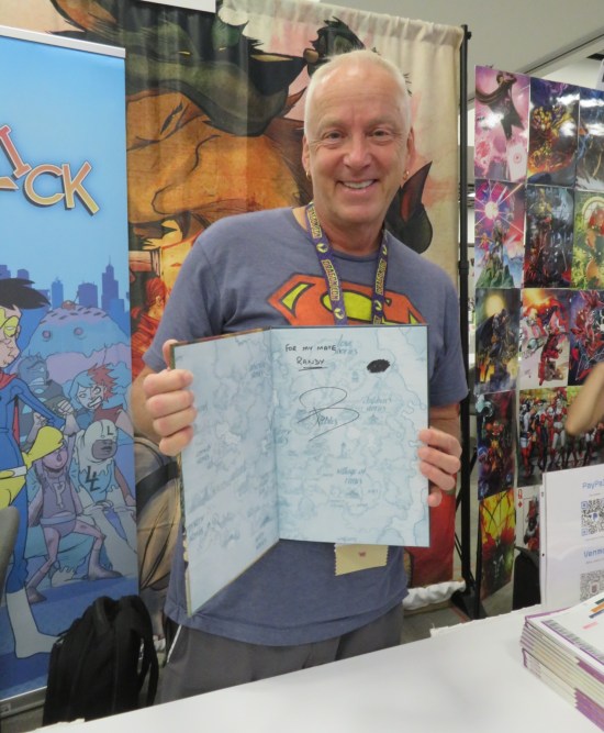 Paul Jenkins holding up a hardcover he just signed and sketched a tiny black cloud with two white eye-holes in it.