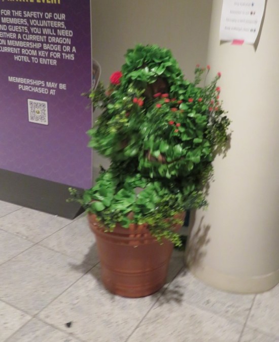 Guy dressed as plant, crouched in giant flowerpot in hallway.