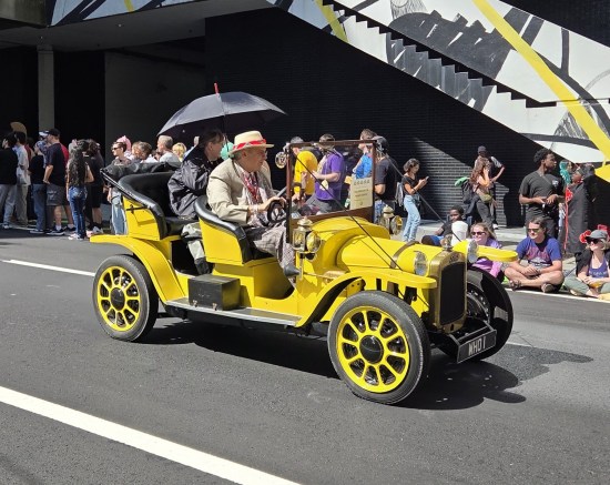 Seventh Doctor cosplayer driving a yellow Edwardian roadster with a darkly dressed passenger crouched behind him.