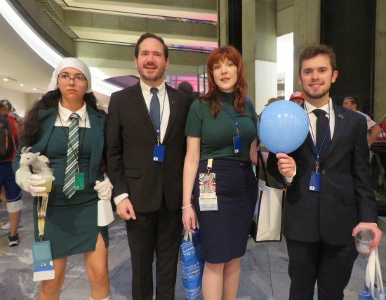 Three people in business suits and one lady in a disheveled green suit a la Gwendolyn Christie's character, holding a stuffed goat.